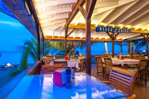 A beachside restaurant with tables and chairs, a sign reading "BEACH & GRILL", and a view of the ocean. - Blue Bahia Resort (Roatan)