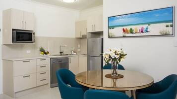 A modern kitchen with white cabinets, a microwave, a dishwasher, and a refrigerator. There is a dining table with blue chairs and a vase with flowers. A beach scene painting is on the wall.