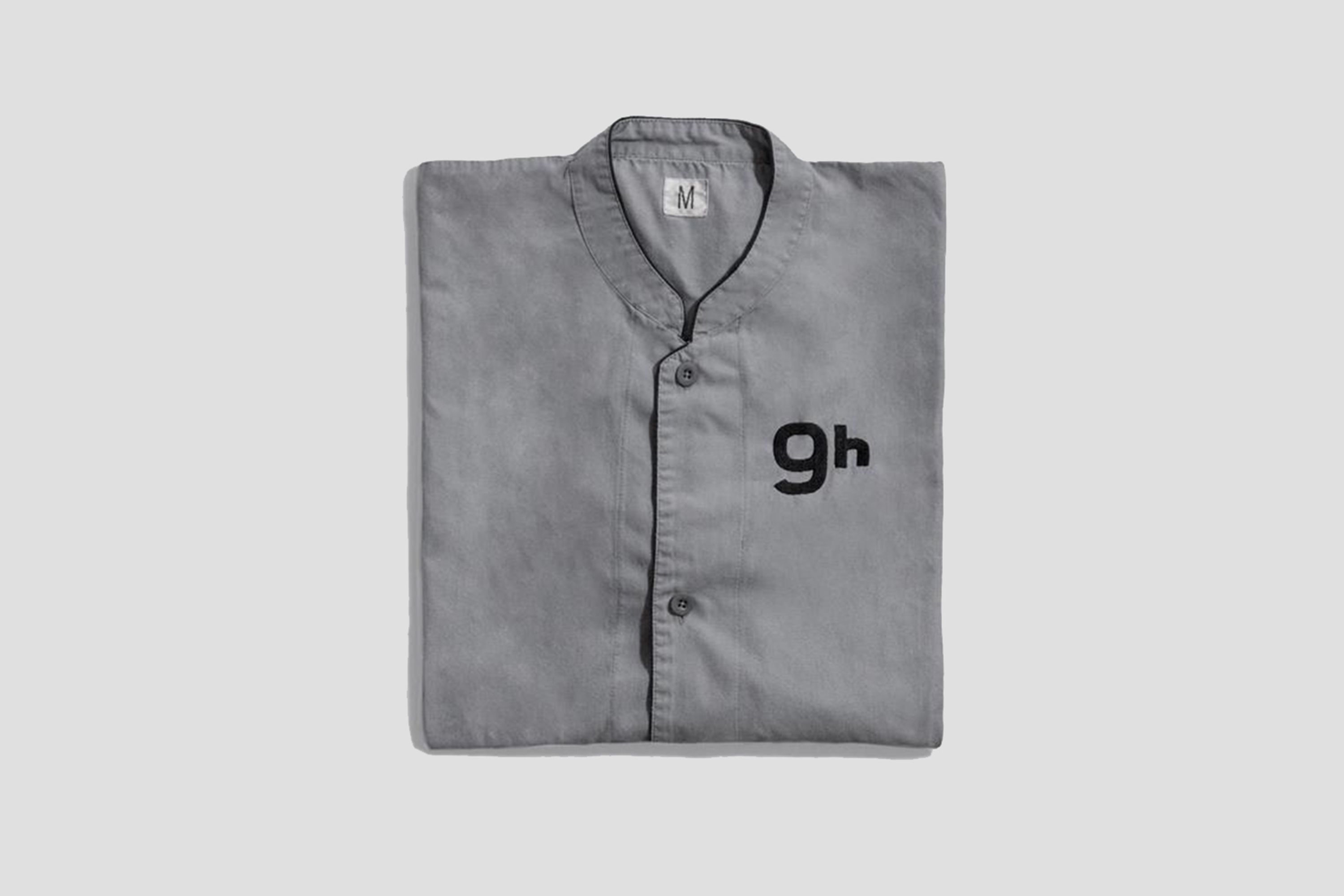 Un t-shirt gris plié, avec un col et des boutons, orné des lettres « gh » sur le devant.