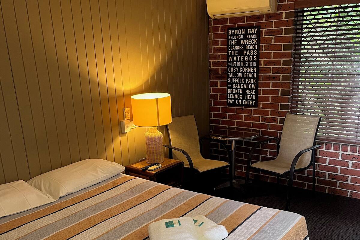 Une chambre d’hôtel avec un lit, une chaise, une table de chevet, une lampe et un mur sur lequel figure une liste de noms de plages.