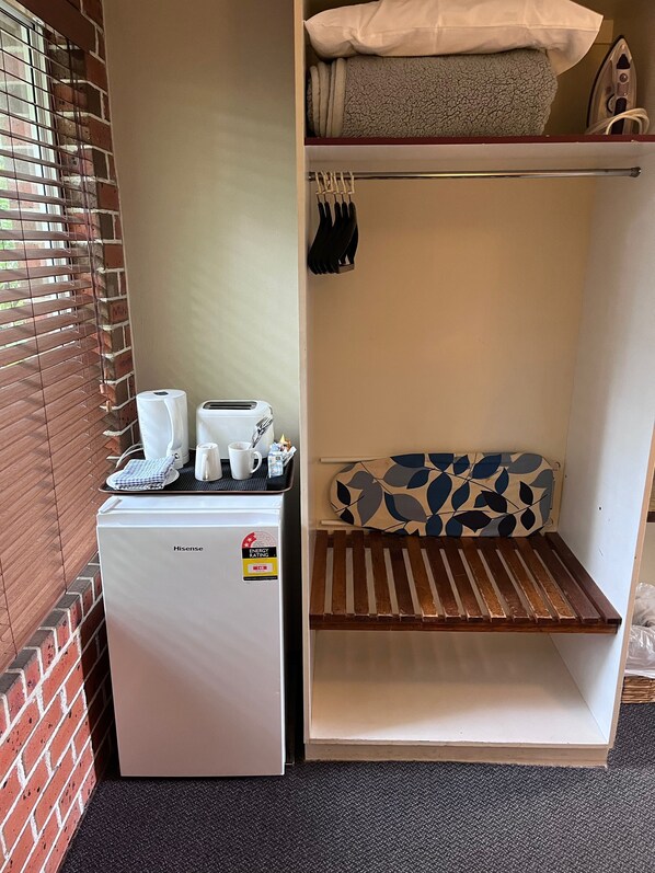 A compact hotel room with a Hisense refrigerator, a small seating area with a patterned cushion, and a shelf with towels and a hairdryer. - The Mullum Motel (Mullumbimby)