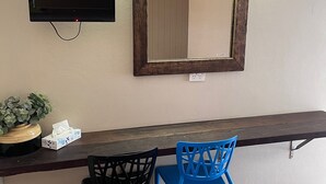 Hotelkamer met een lang houten bureau, twee stoelen, een televisie, een spiegel, een kleine plant en een afvalbak.