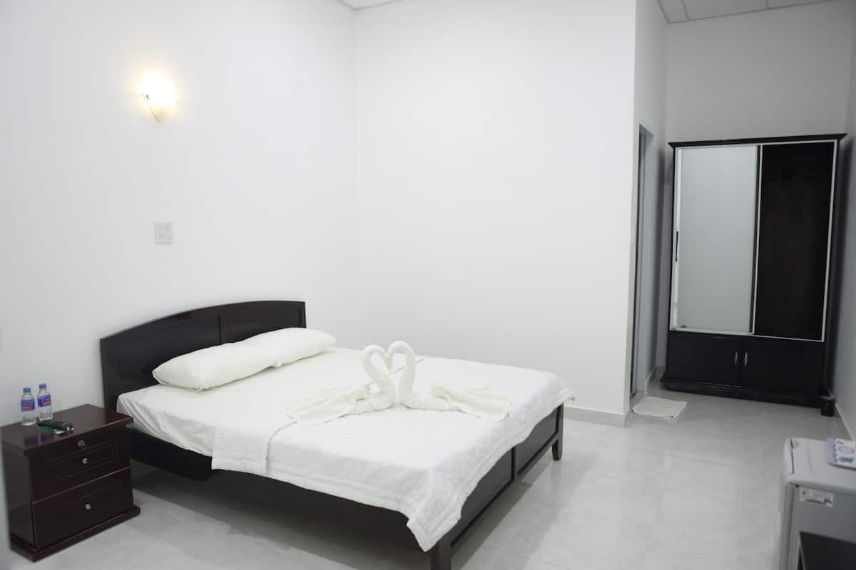 A neatly made bed with white linens and a heart-shaped towel arrangement, a dark wooden nightstand with a remote control and water bottles, and a large mirror on the wall.