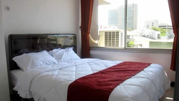 A neatly made bed with white bedding and a red blanket, a window with curtains, and a view of buildings outside. - Providence Homestay (Surabaya)