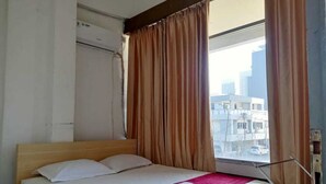 A hotel room with a bed, a window with curtains, and a view of buildings outside. - Providence Homestay (Surabaya)