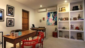 A dining area with a wooden table and red chairs, a bookshelf with various items, and a large abstract painting on the wall. - The Apartment Umalas (Keroboken Kelod)