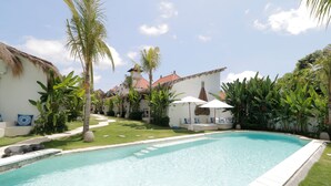 A swimming pool with a thatched-roof building in the background, surrounded by lush greenery and a clear sky. - The Apartment Umalas (Keroboken Kelod)