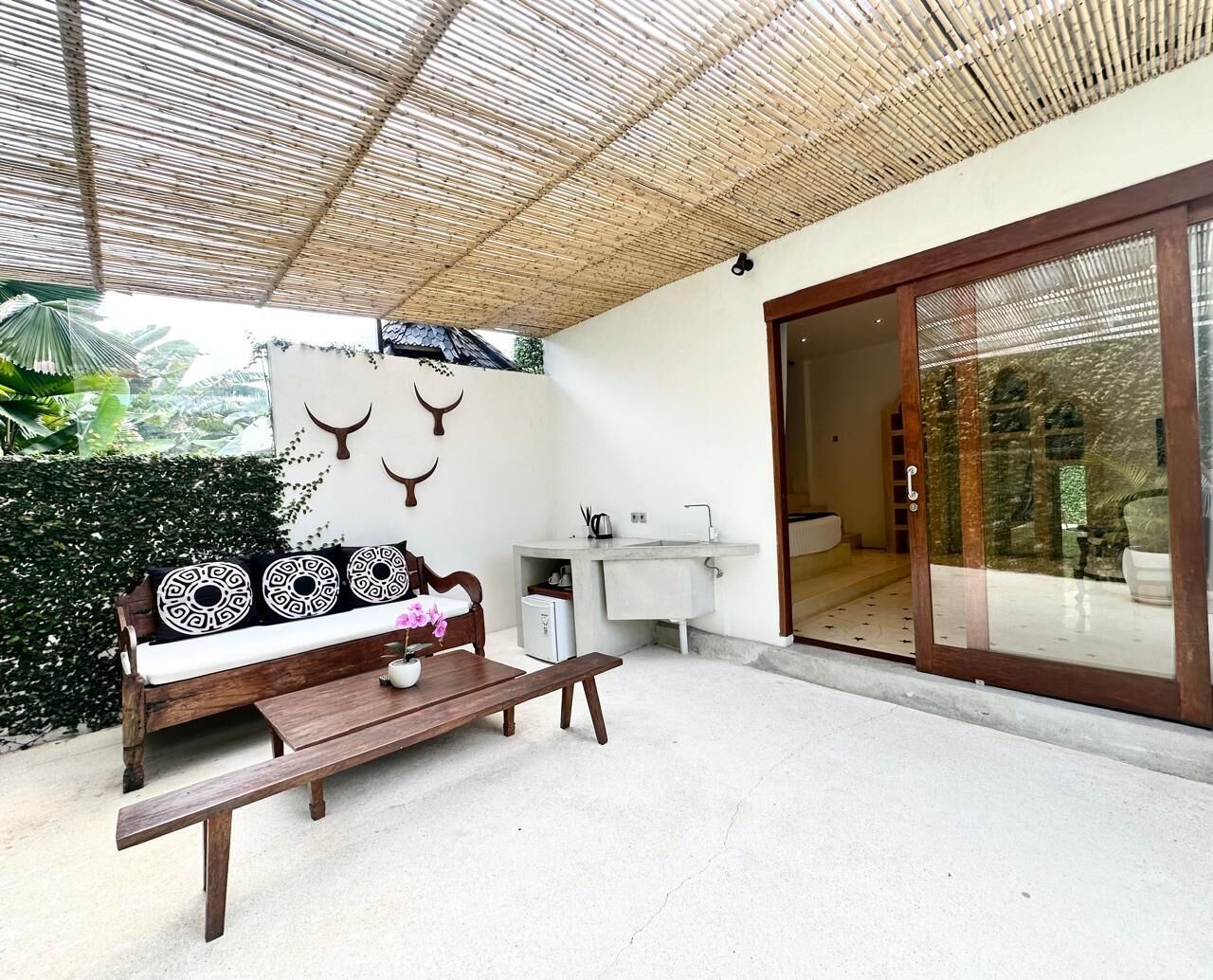 A covered outdoor seating area with a wooden bench and a sofa, a small table, a sink, and a sliding glass door leading to an interior space.