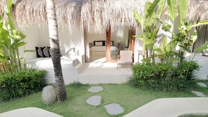 A tropical resort room with a thatched roof, a bed, a chair, and a small garden area with a pathway. - The Apartment Umalas (Keroboken Kelod)