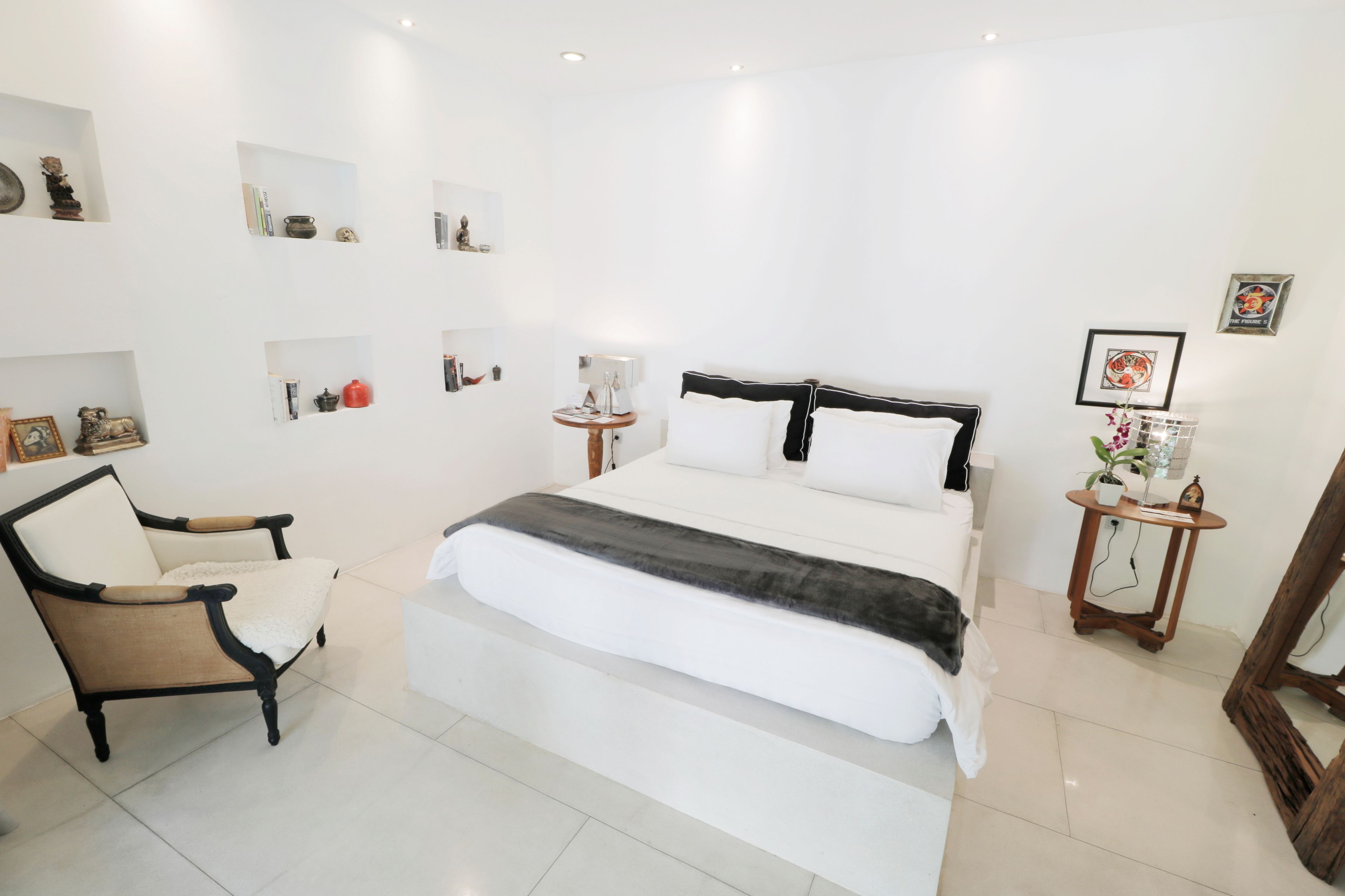A bedroom with a large bed, a chair, a bedside table, and shelves with various items.