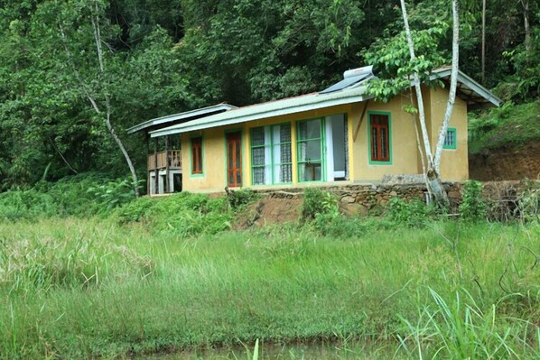 A single-story house with a stone foundation, surrounded by lush greenery and a small body of water.