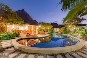 A circular pool with a stone border, surrounded by lounge chairs and a thatched-roof building.