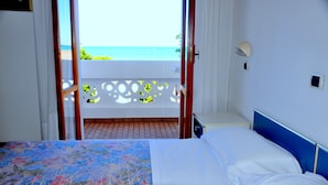 Quarto de hotel com cama, mesa de cabeceira pequena, luminária na parede e vista para o oceano através de uma varanda com tela decorativa.