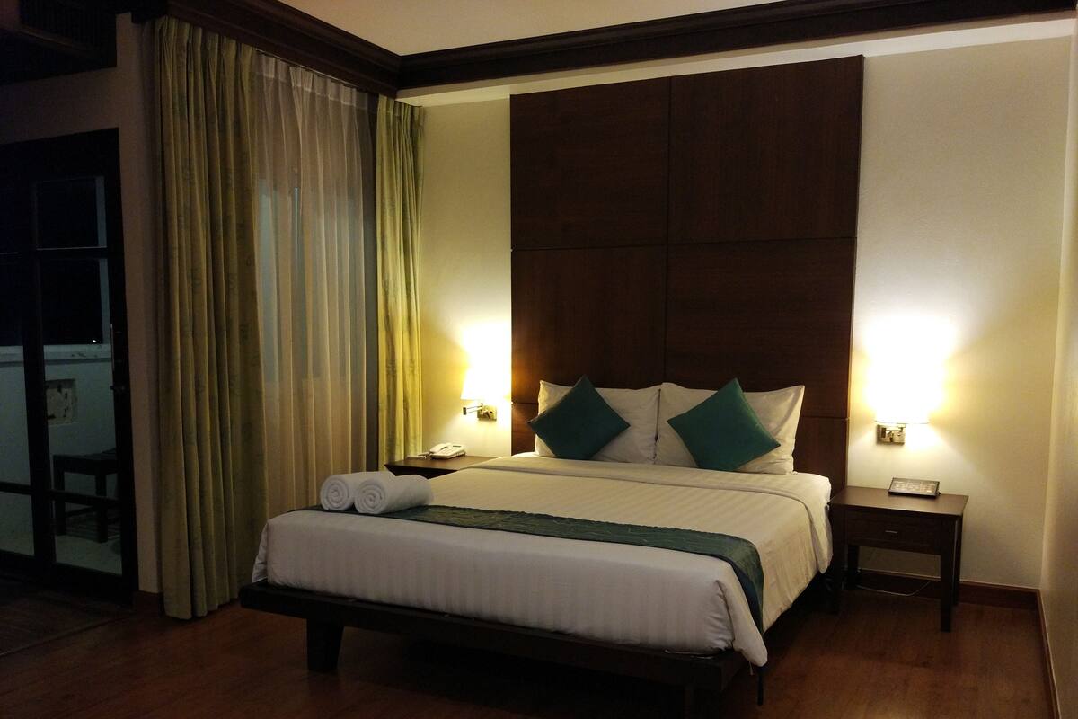 A hotel room with a large bed, two bedside lamps, a wooden headboard, a nightstand, and a window with curtains.