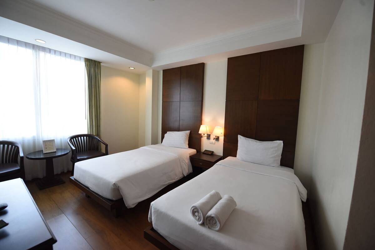 A hotel room with two beds, a small table, two chairs, a desk, and a window with curtains.