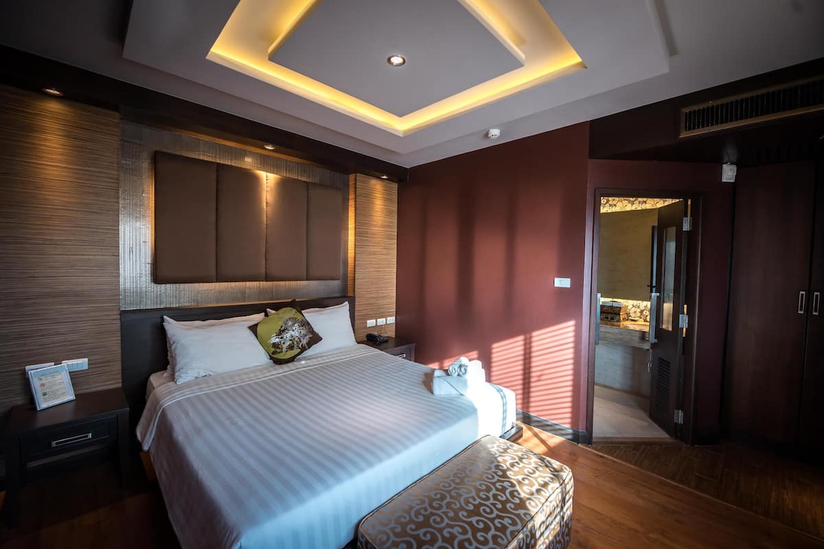 A modern hotel room with a large bed, a bedside table, a nightstand, a patterned ottoman, and a ceiling light fixture.