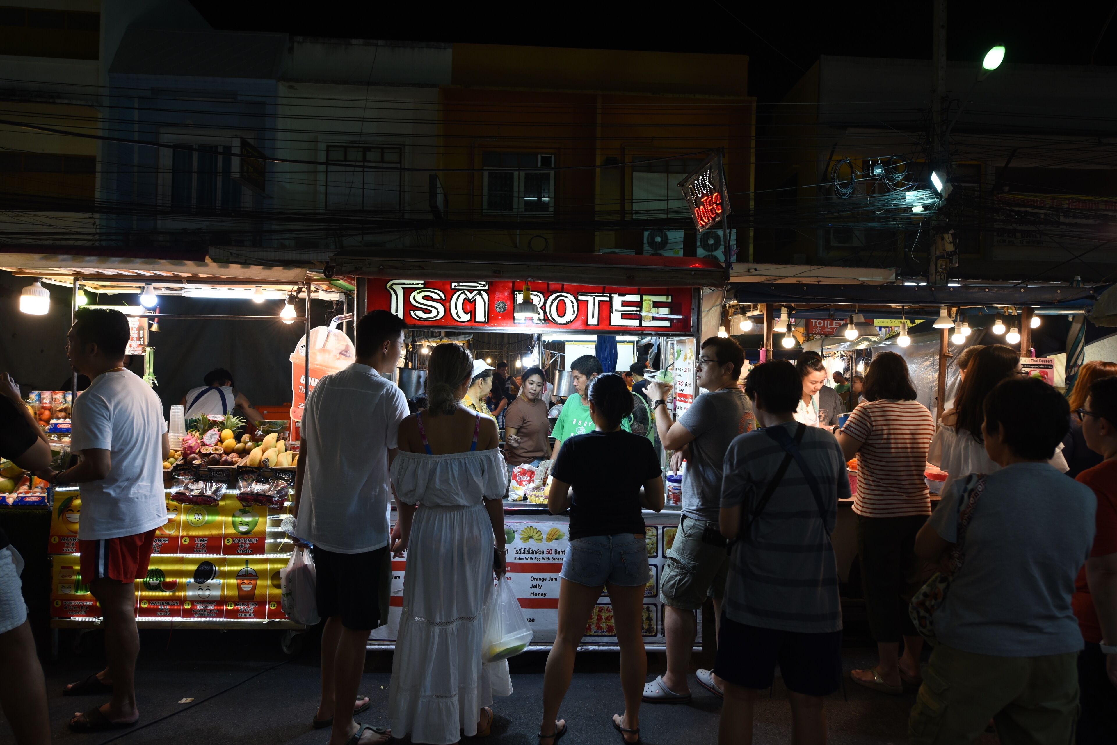 Un puesto de comida callejera concurrido con un letrero de neón, rodeado de gente.