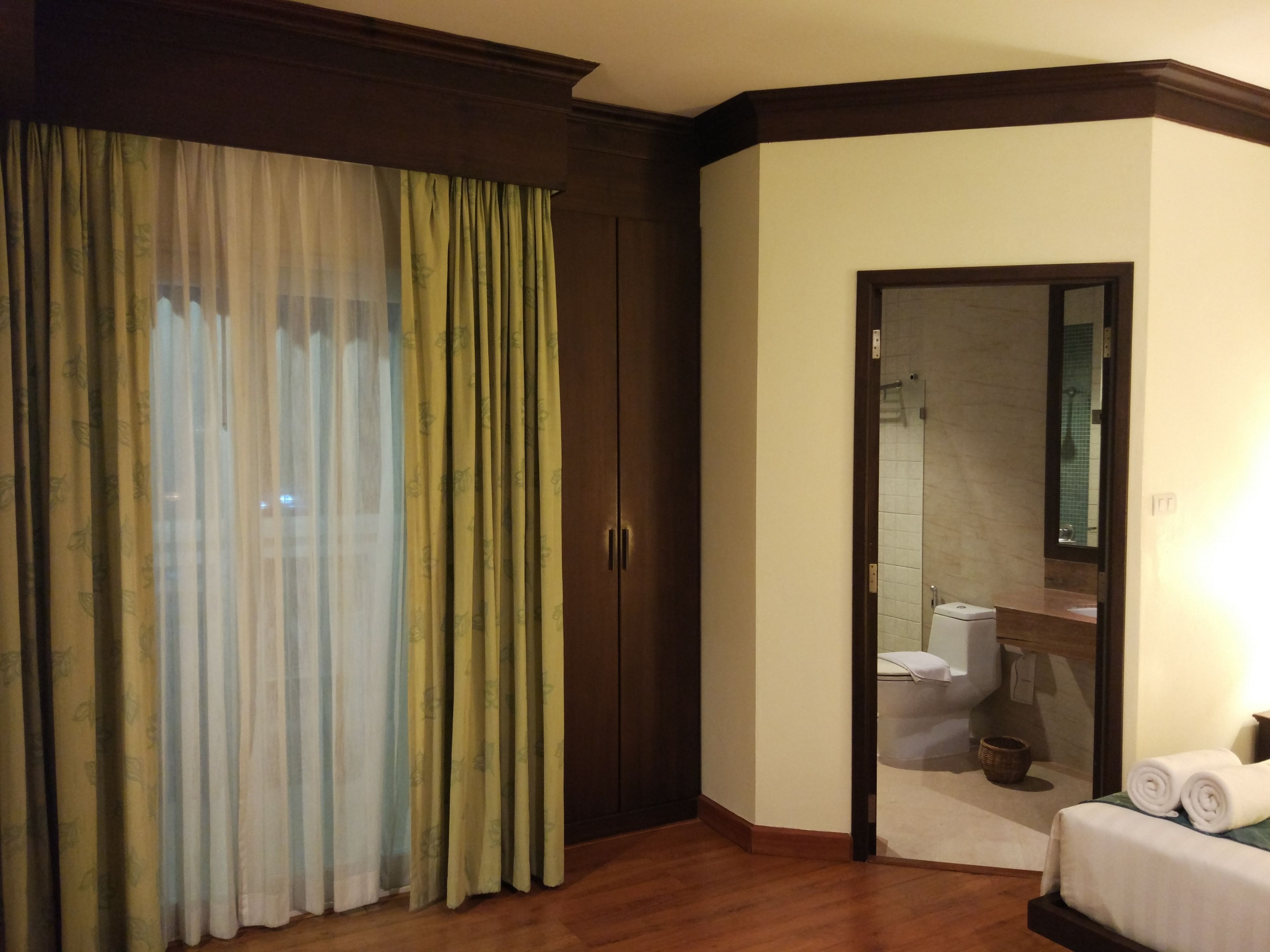 A hotel room with a wooden floor, a large window with sheer curtains, a wardrobe, a bathroom with a mirror, and a bed with rolled towels.