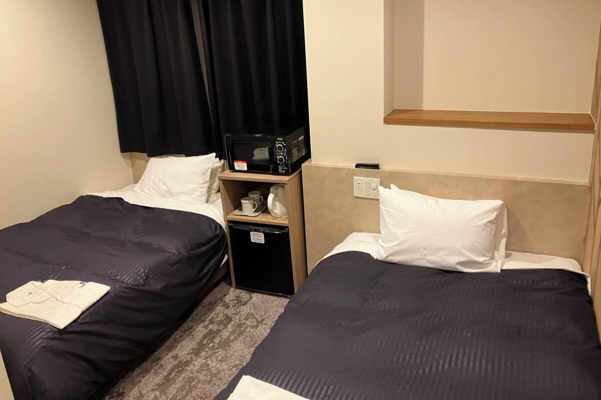 A hotel room with two beds, a microwave, a small table with a cup and a kettle, and a shelf above the bed.