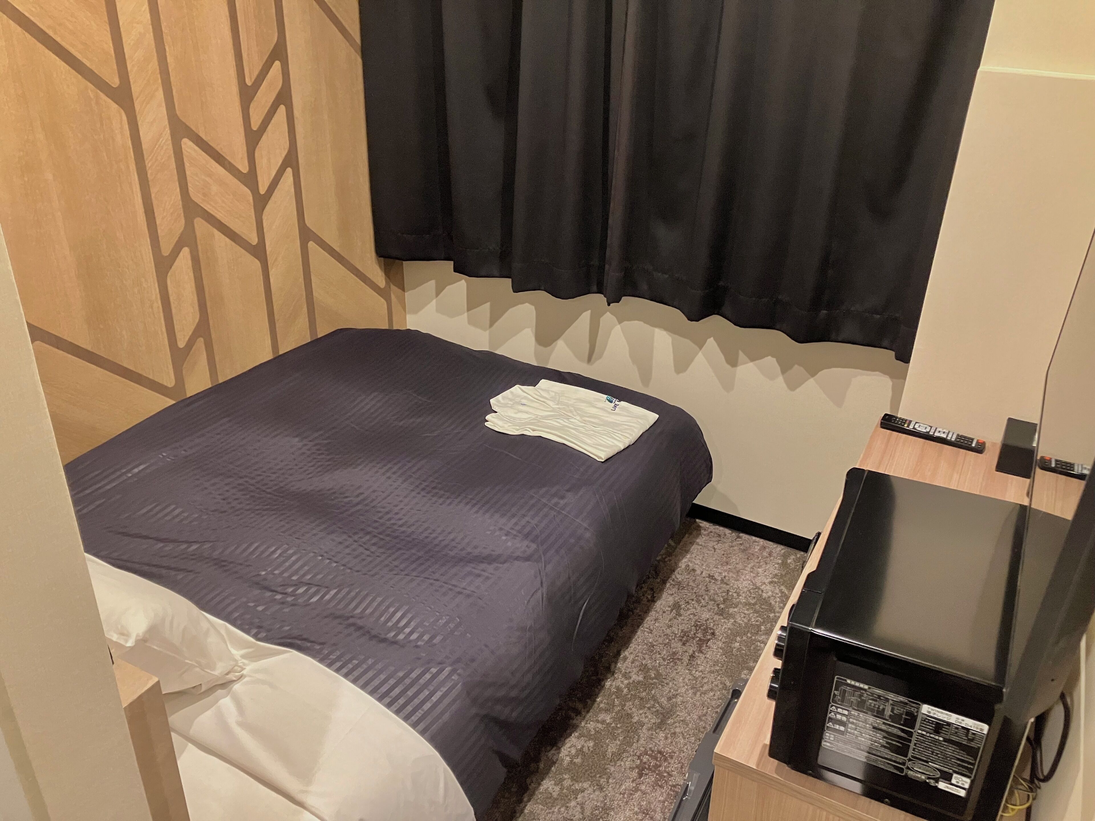 A small, neatly made bed with a dark blue bedspread and a white pillow. There is a remote control and a small black device on a wooden nightstand beside the bed.