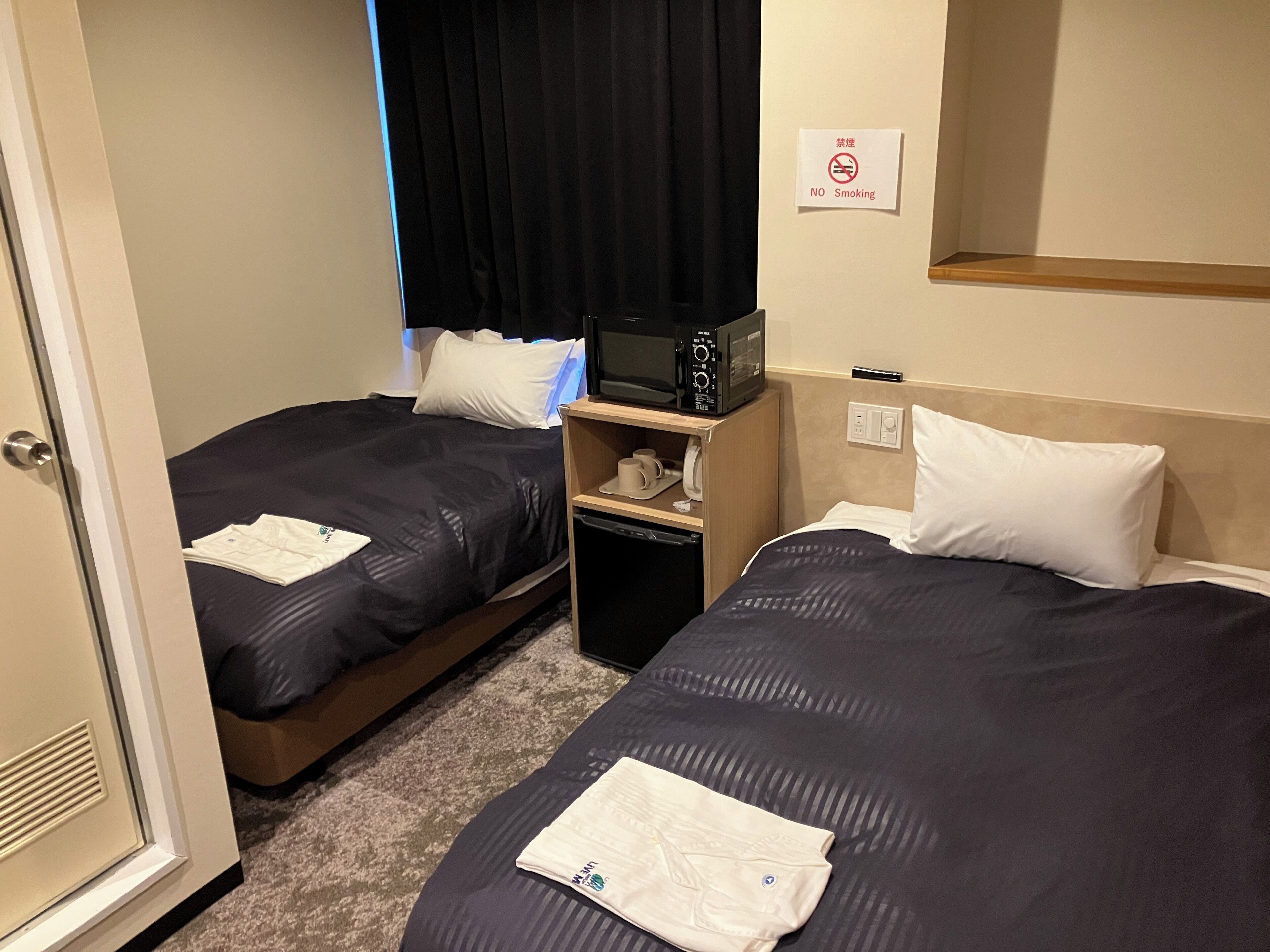 Habitación de hotel con dos camas, un microondas, un estante pequeño con artículos de aseo y un cartel de prohibición de fumar.