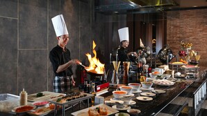 Een kok bereidt eten met vuur op een fornuis in een moderne keuken, met een buffet vol diverse gerechten.