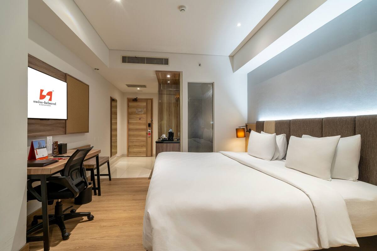Una habitación de hotel con cama, escritorio, sillas y televisor mostrando el nombre del hotel.