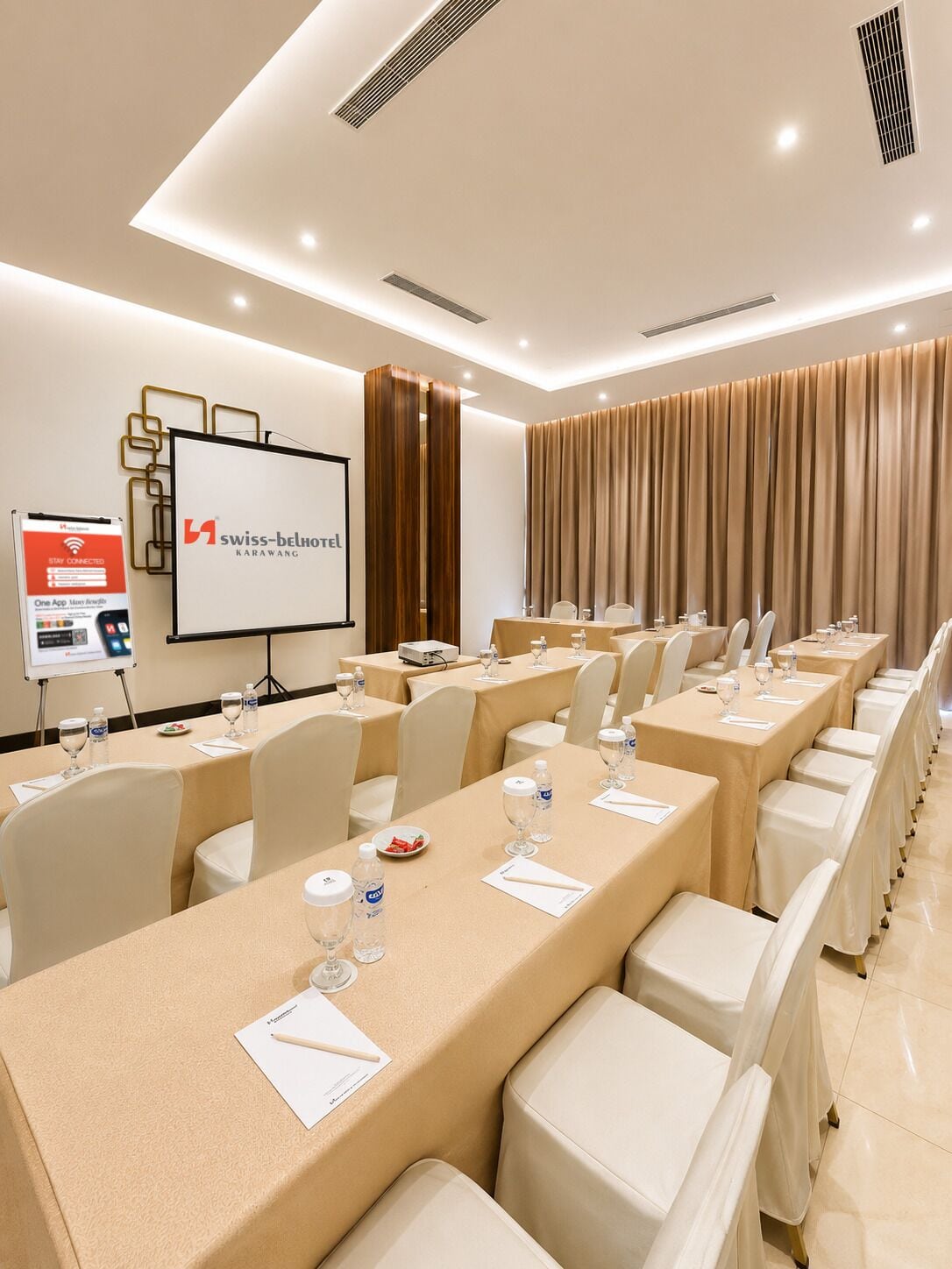 Un salón de conferencias con mesas dispuestas para una reunión, una pantalla de proyector y un letrero de Swiss-Belhotel Karawang.