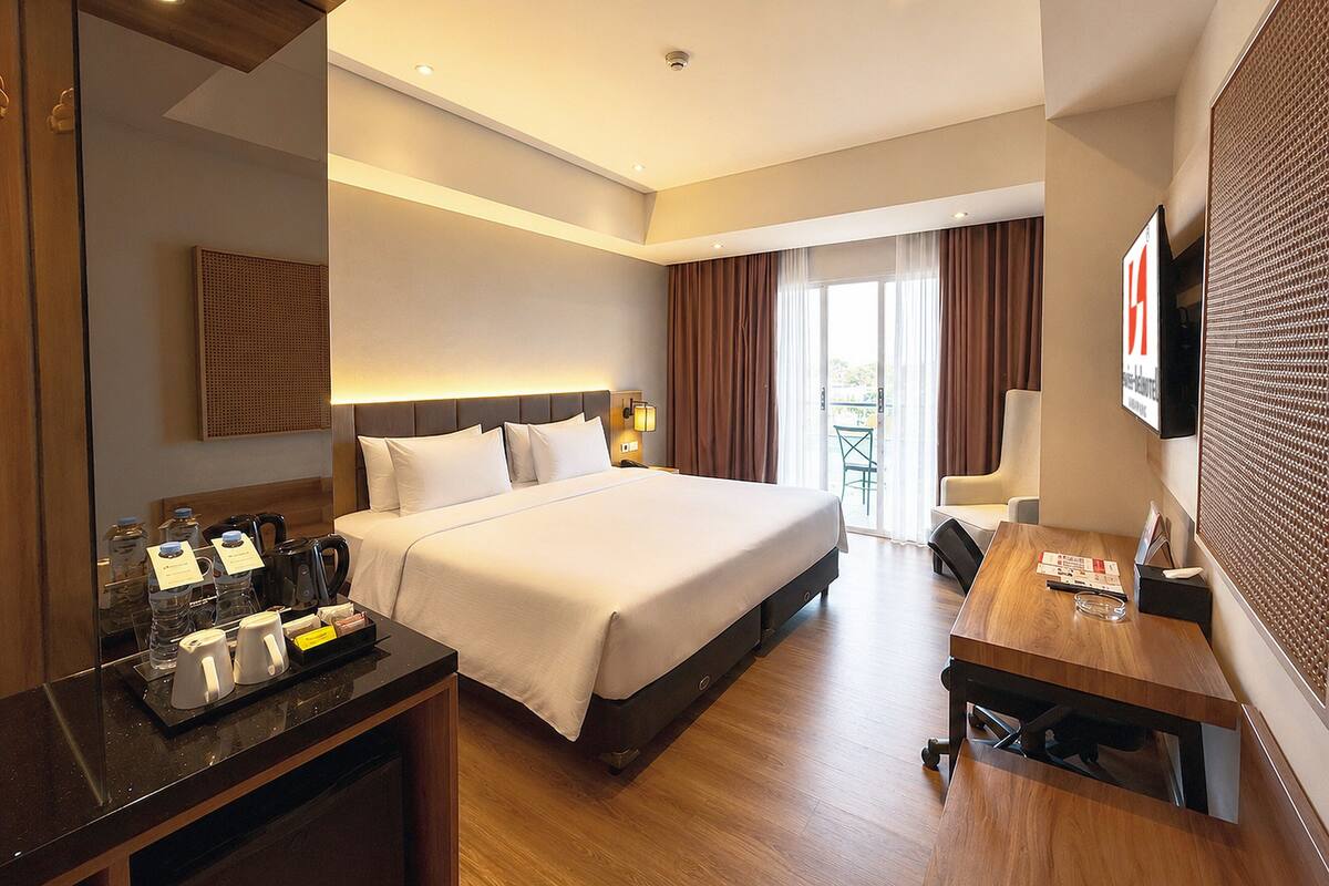 Habitación de hotel con cama, escritorio, silla, televisor y balcón con cortinas.