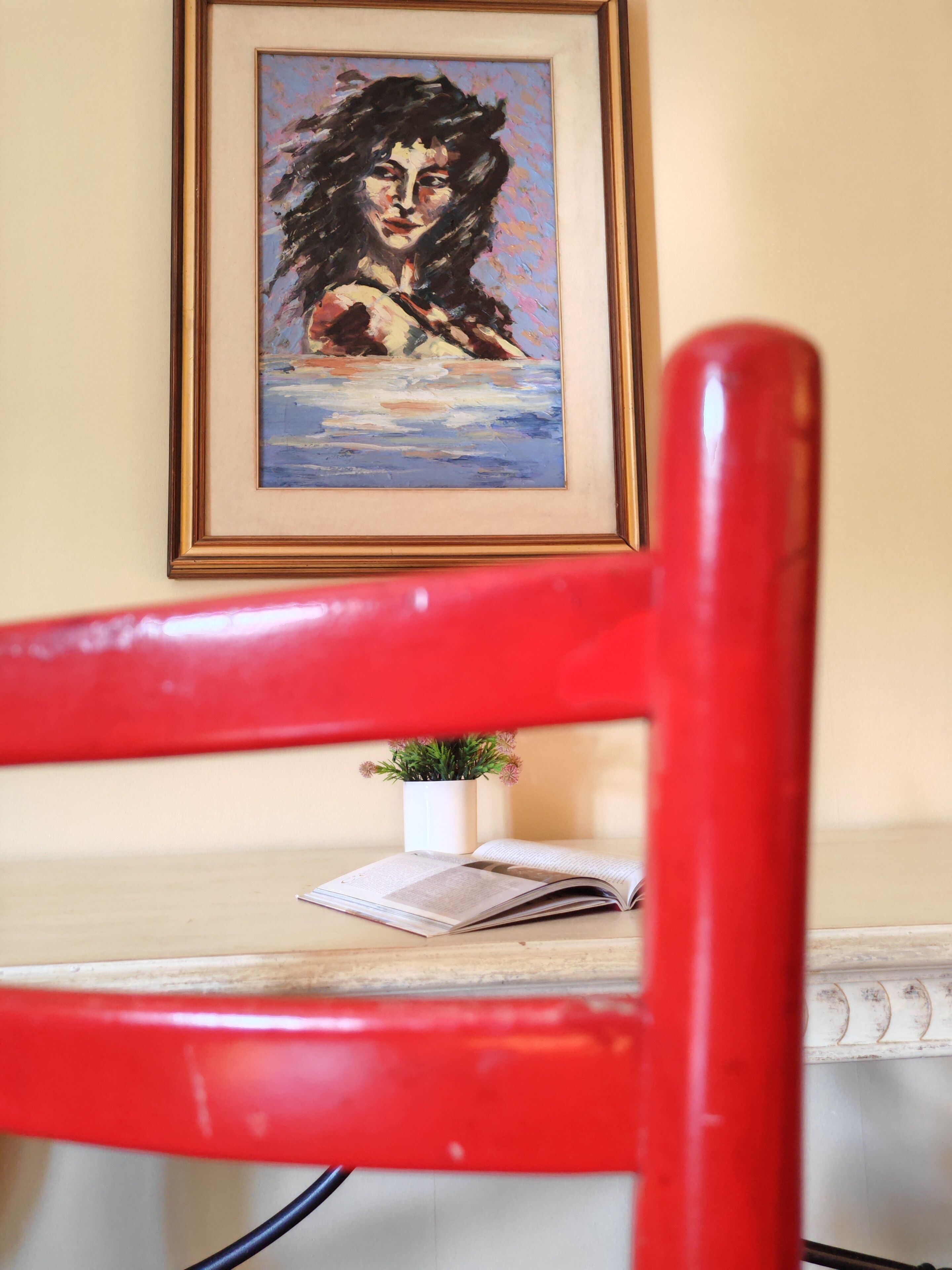 A red chair in front of a table with a framed painting, a small potted plant, and an open book.