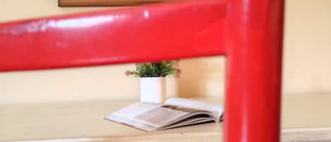 A red chair in front of a table with a framed painting, a small potted plant, and an open book.