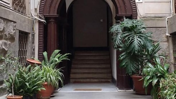 Entrada de un edificio con un arco imponente, escalones que conducen a una puerta y macetas con plantas a cada lado.