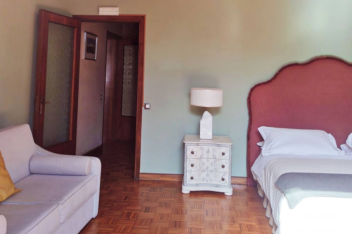 A room with a bed, a sofa, a nightstand, a lamp, and a door leading to another room.