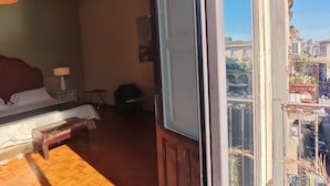 Una habitación con piso de madera, una cama con ropa blanca, una mesita de noche con lámpara y vistas a un paisaje urbano a través de un ventanal grande.