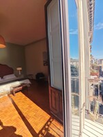 Una habitación con piso de madera, una cama con ropa blanca, una mesita de noche con lámpara y vistas a un paisaje urbano a través de un ventanal grande.