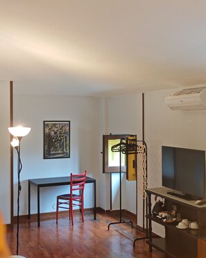 A room with a wooden floor, a desk with a chair, a TV on a stand, a lamp, and a painting on the wall. - Liodoro Bed and Breakfast (Catania)