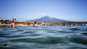 Una playa con gente nadando y tomando el sol, una montaña al fondo y un cielo azul despejado.