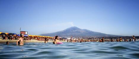 A beach with people swimming and sunbathing, a mountain in the background, and a clear blue sky.