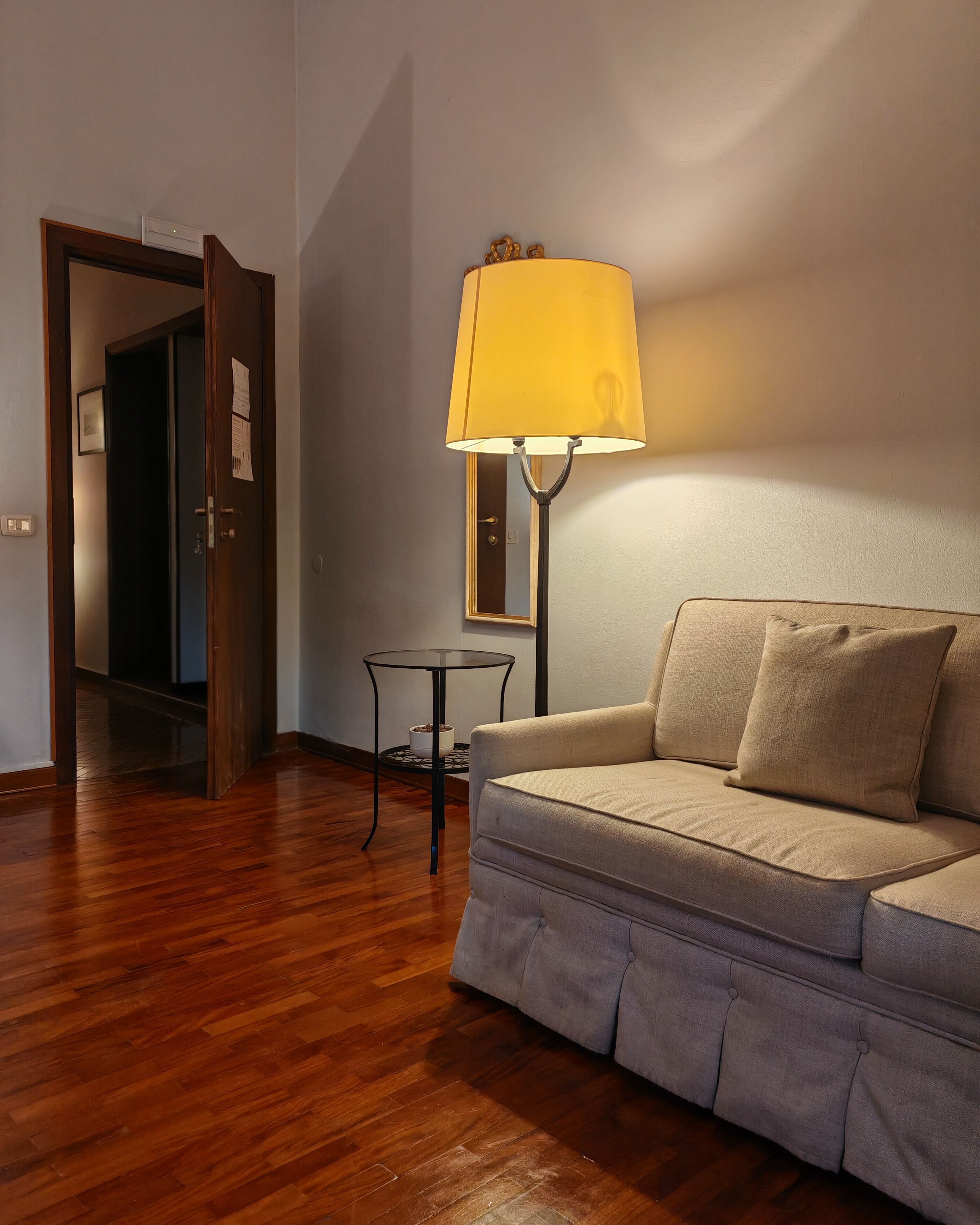 Une pièce avec un plancher en bois, un canapé beige, une petite table ronde avec une lampe, et une porte qui donne sur une autre pièce.