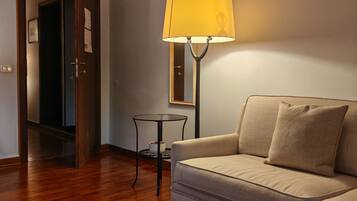 Une pièce avec un plancher en bois, un canapé beige, une petite table ronde avec une lampe, et une porte qui donne sur une autre pièce.