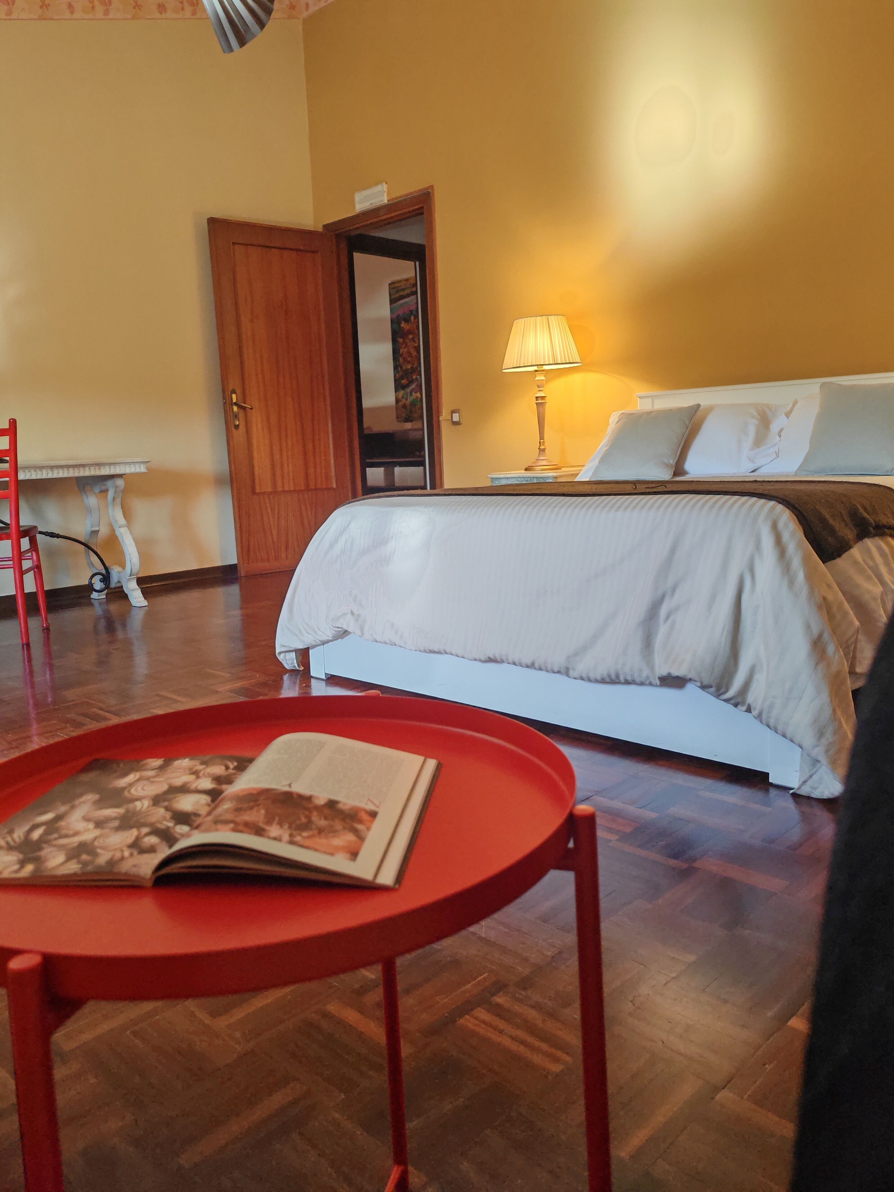 Une chambre d’hôtel avec un lit, une table de chevet avec une lampe, une chaise rouge et une petite table avec un livre.