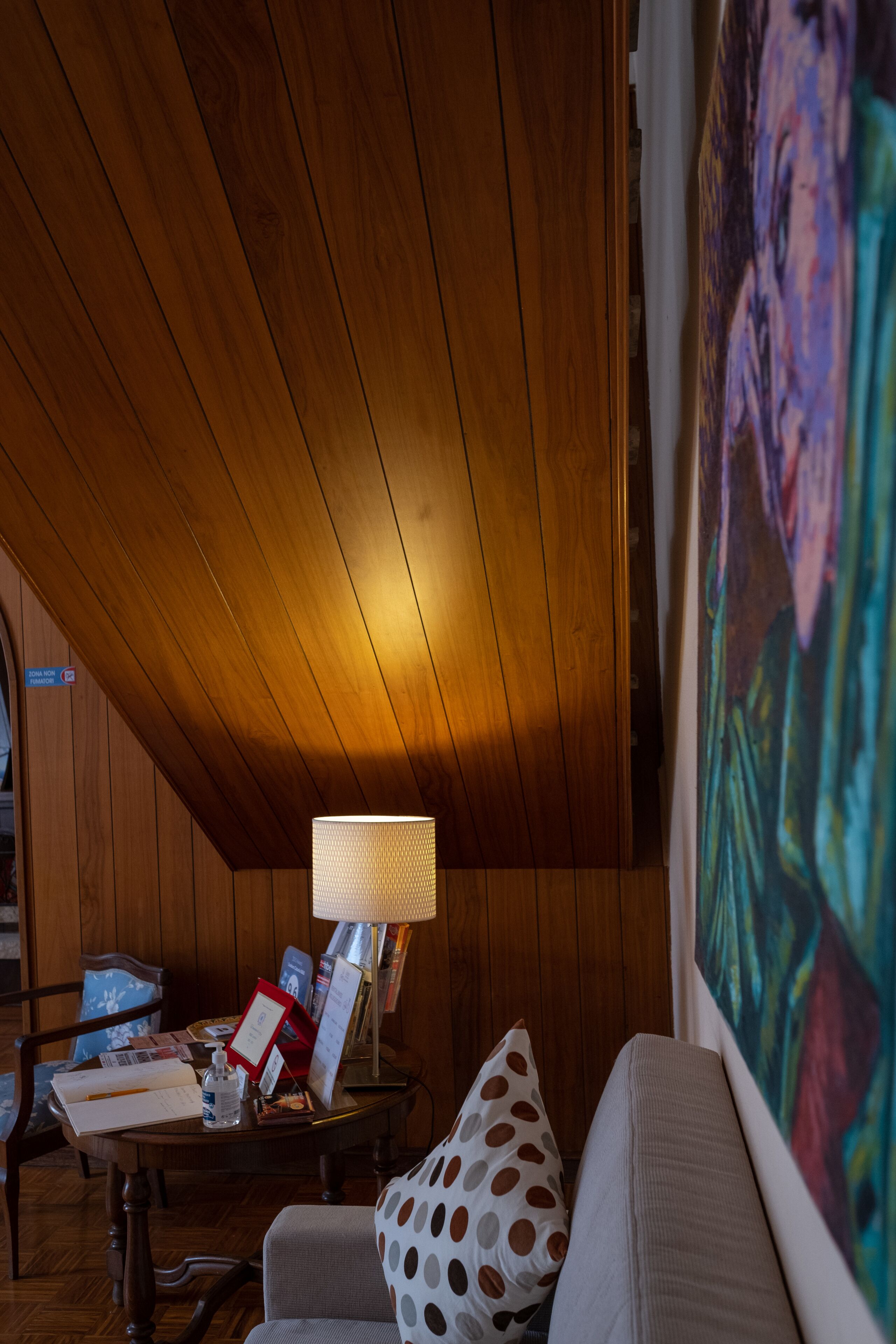 Una habitación acogedora con paneles de madera, una mesa redonda con lámpara, un sofá con un cojín de lunares y un cuadro abstracto de gran tamaño.