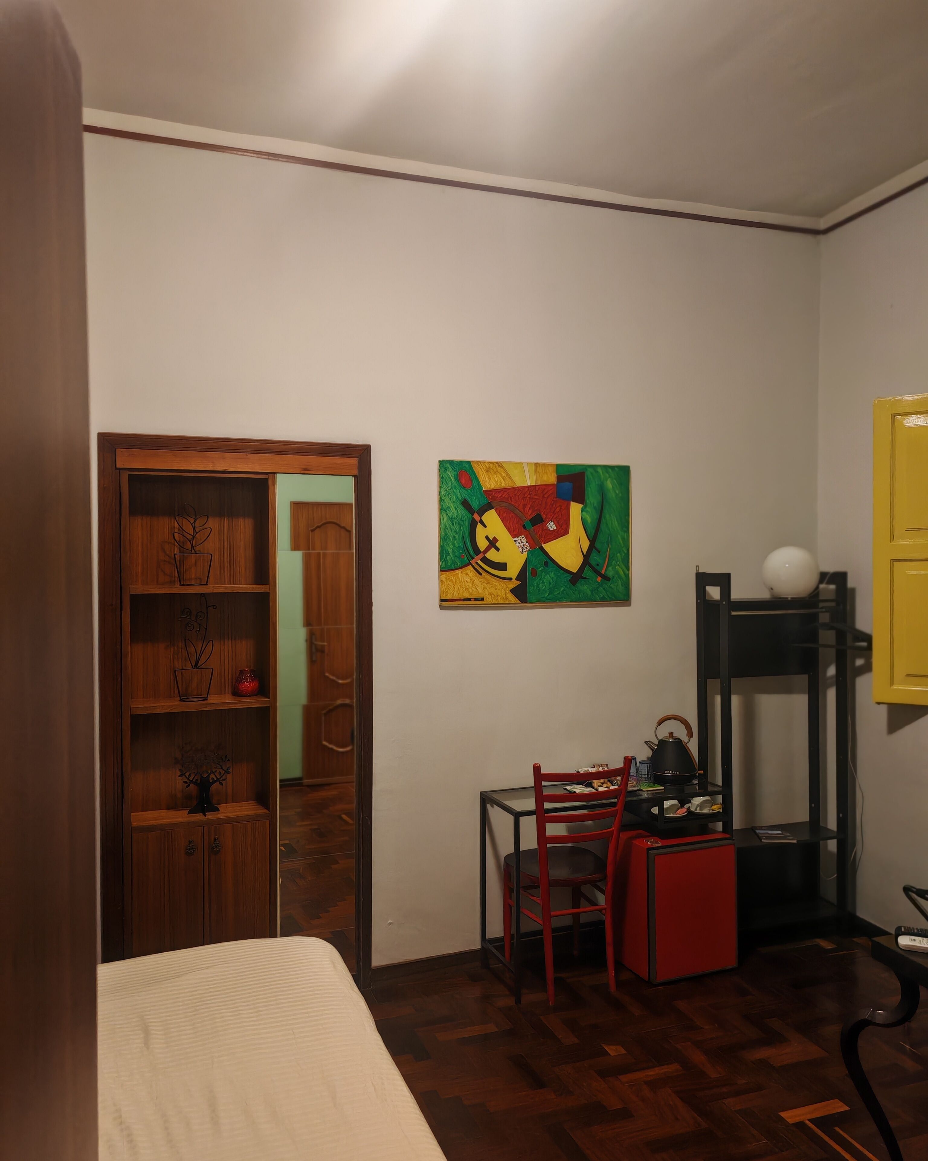 Een kamer met een bed, een houten kledingkast, een rode stoel, een klein tafeltje met een waterkoker, en een kleurrijk abstract schilderij aan de muur.