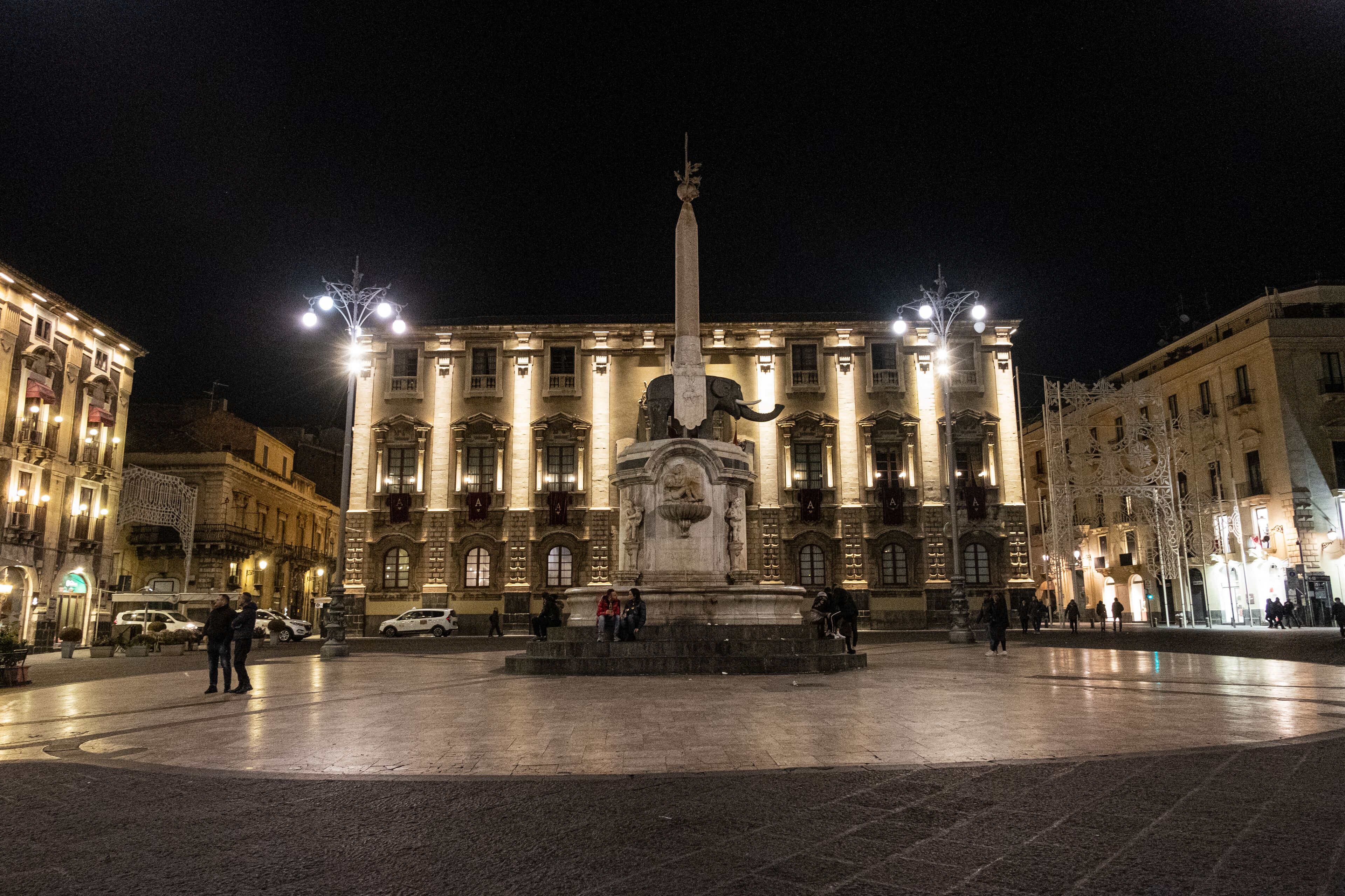 Una plaza con un monumento central, rodeada de edificios históricos con fachadas iluminadas.