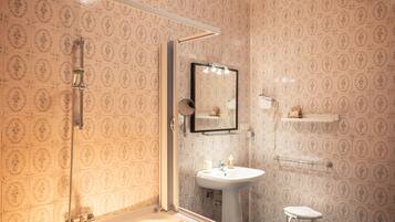 A bathroom with patterned tiles, a pedestal sink, a mirror, a shower, and a small stool.
