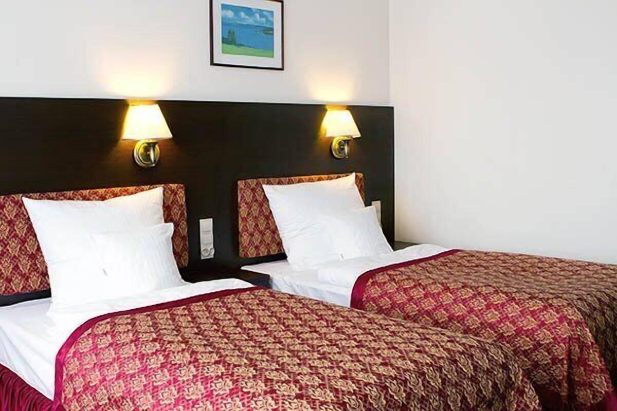 A hotel room with two beds, each with a patterned bedspread and white pillows. There are two wall-mounted lamps on either side of the beds.