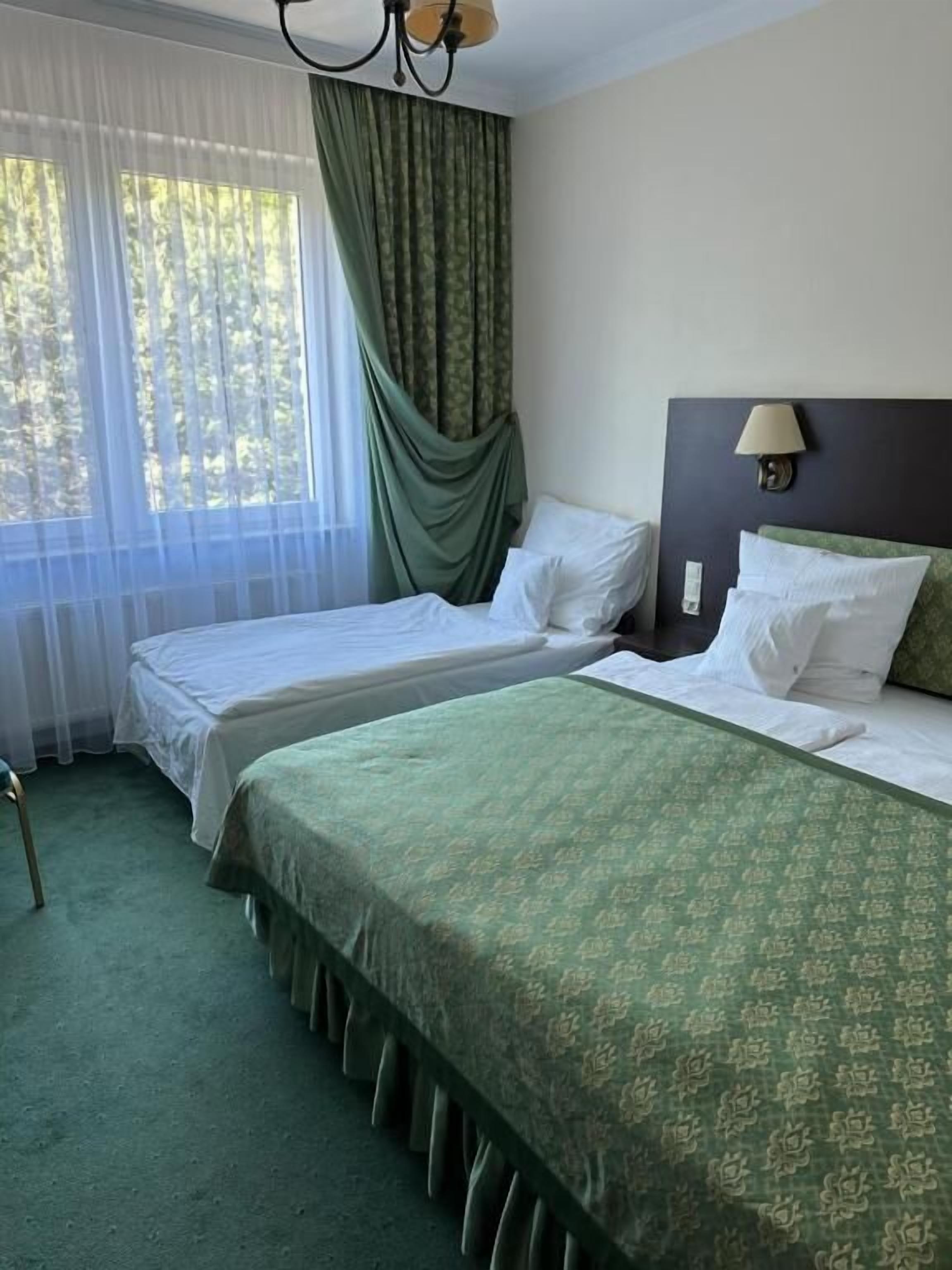 Habitación de hotel con dos camas, una ventana con cortinas translúcidas, un cabecero con lámpara y alfombra verde.