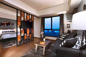 A modern hotel room with a bed, a sofa, a coffee table, and a large window offering a view of the ocean.