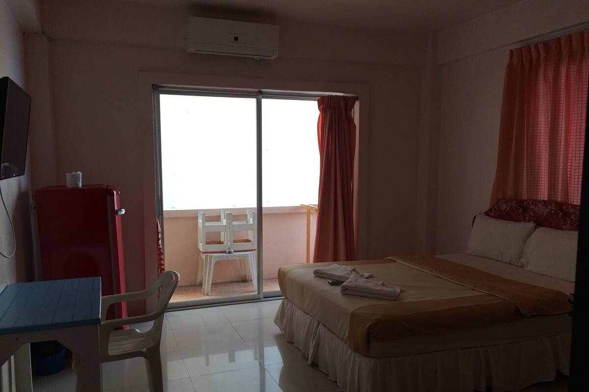 Habitación de hotel con cama, escritorio, silla, refrigerador rojo y puerta corrediza de vidrio que da a un balcón.