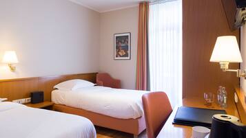 A hotel room with two beds, a desk with a lamp, a picture on the wall, and a window with curtains.
